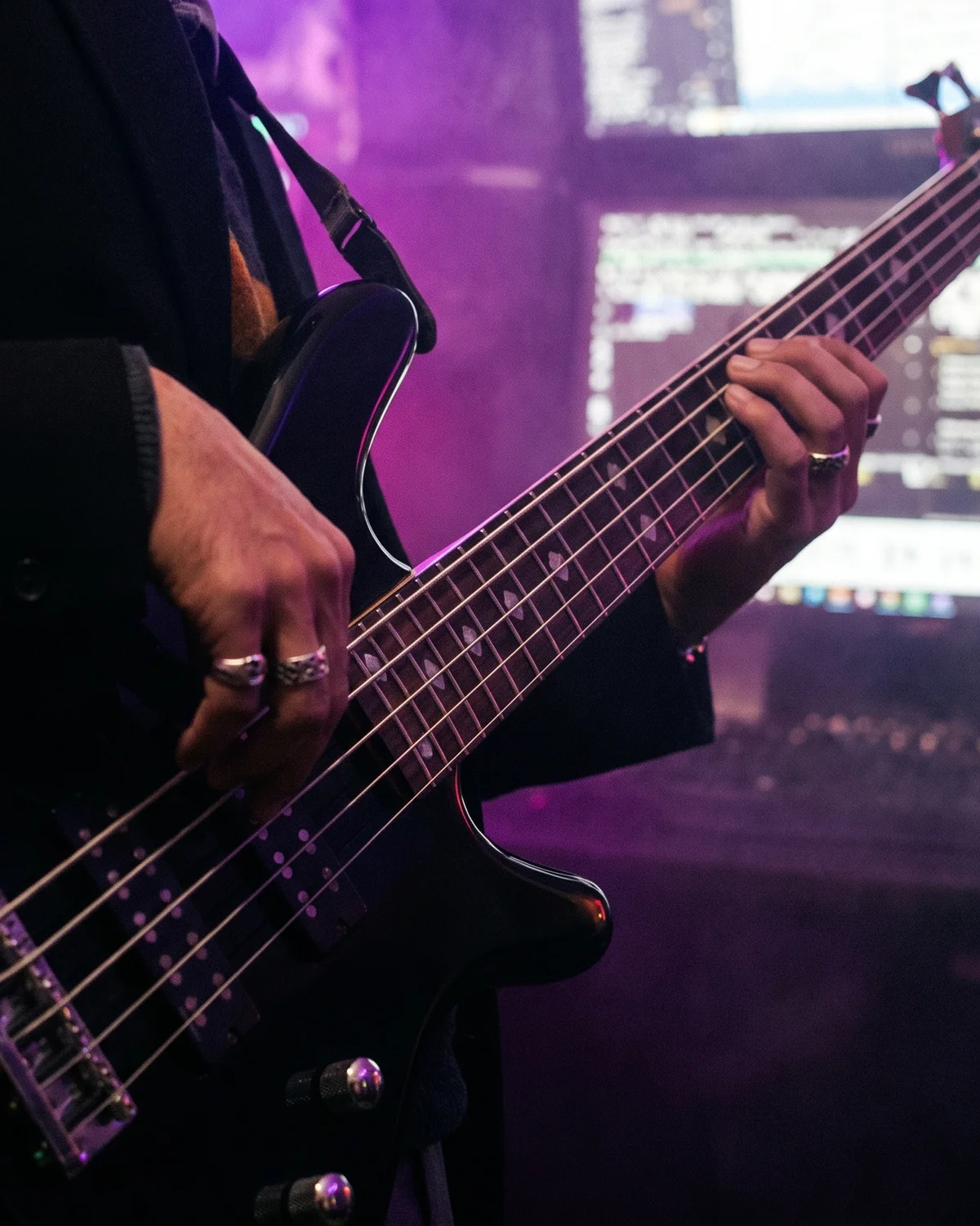 Hands playing a black five-string bass guitar in a purple-lit home studio